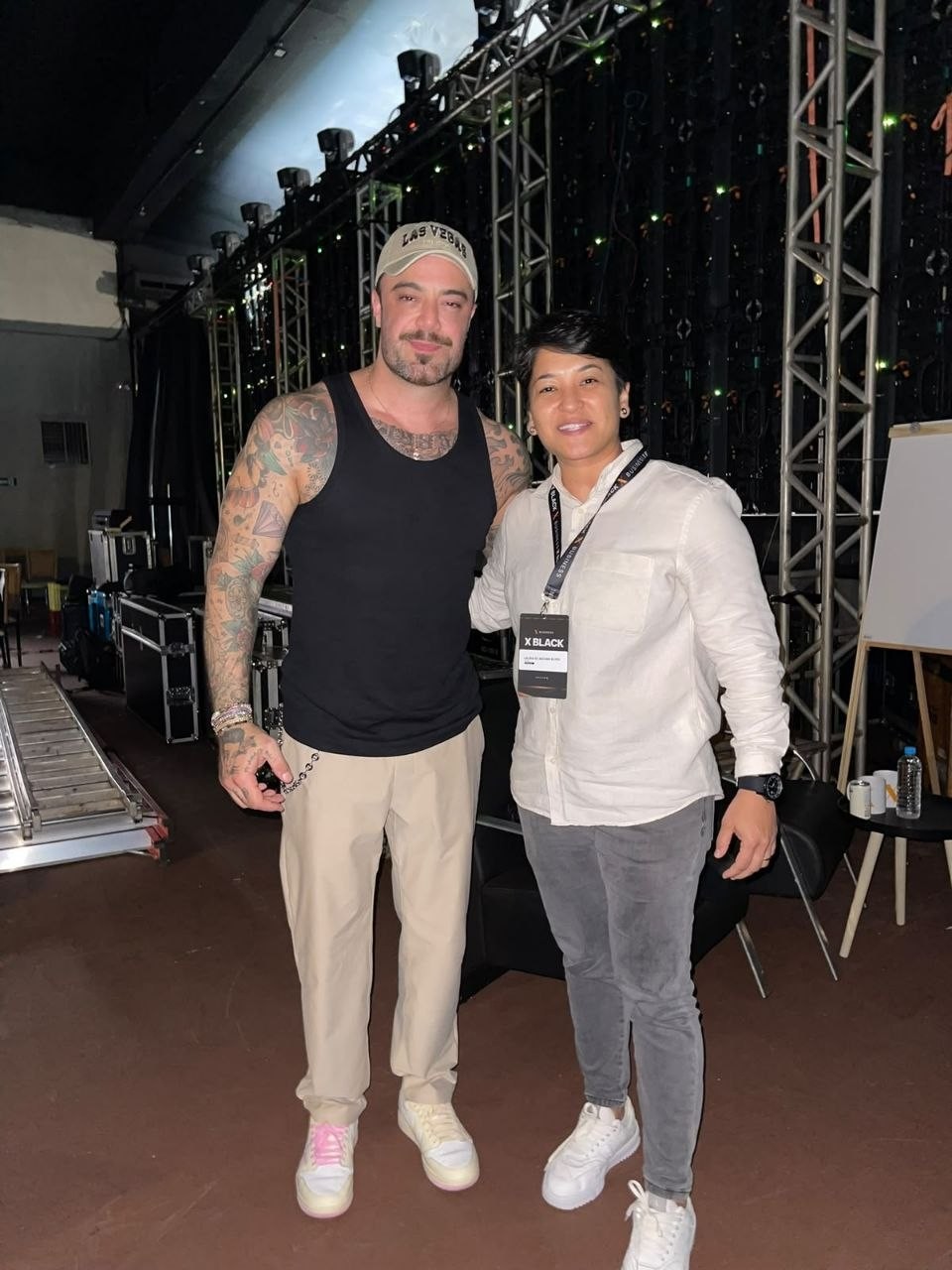 Felipe Titto e Laura Alves posam lado a lado nos bastidores de um evento, com estrutura de palco e iluminação ao fundo